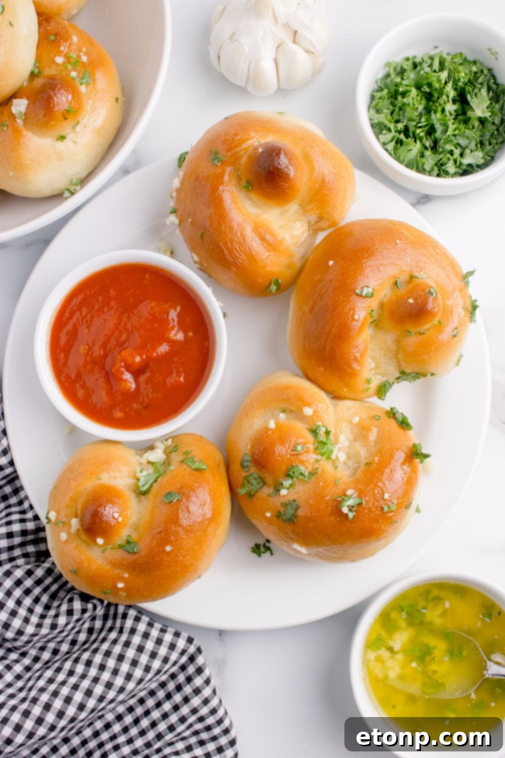 Homemade Garlic Knots spread out on plate with some marinara on it, and a side of garlic butter sitting on table beside the bowl 