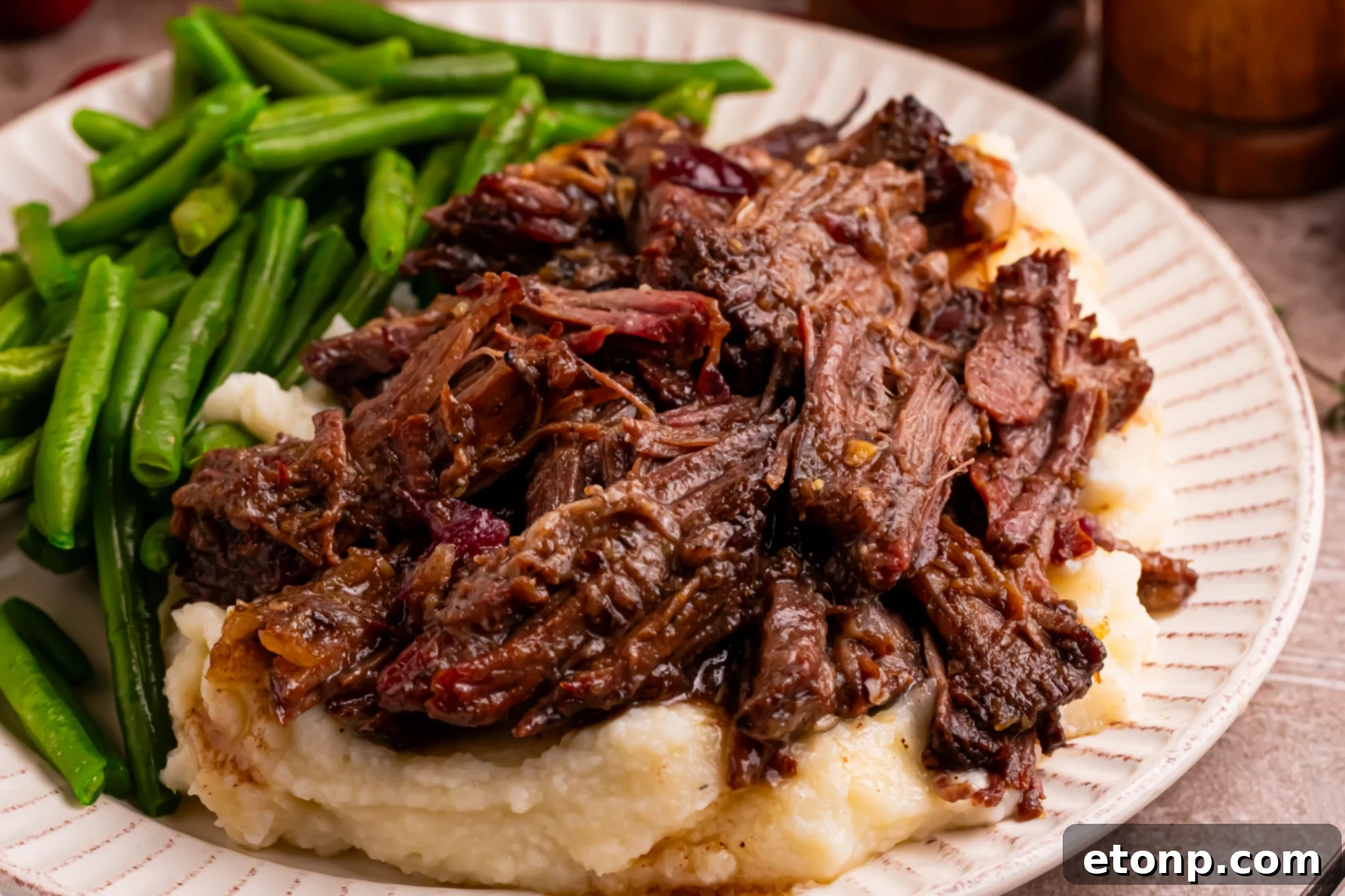 Juicy Cranberry Pot Roast shredded on a white plate with mashed potatoes under and a side of green beans 