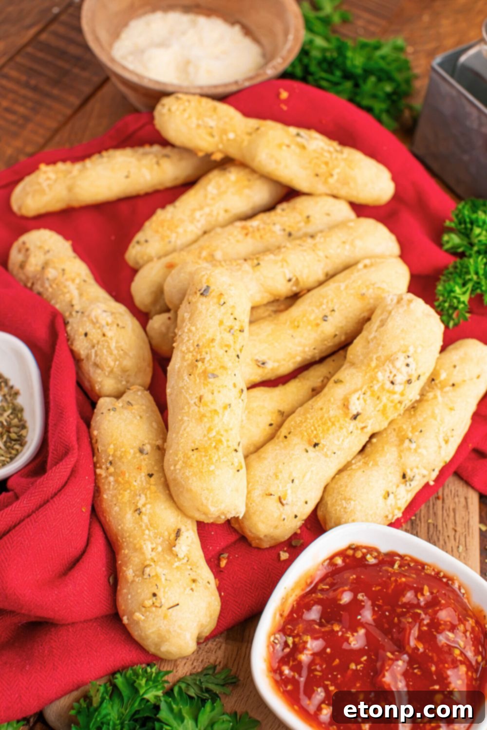 Easy Buttery Garlic Parmesan Breadsticks Two Ingredients 13 A red kitchen towel laid on a table, with freshly baked 2-Ingredient Garlic Parmesan Breadsticks scattered around, and a bowl of dipping sauce to the side.