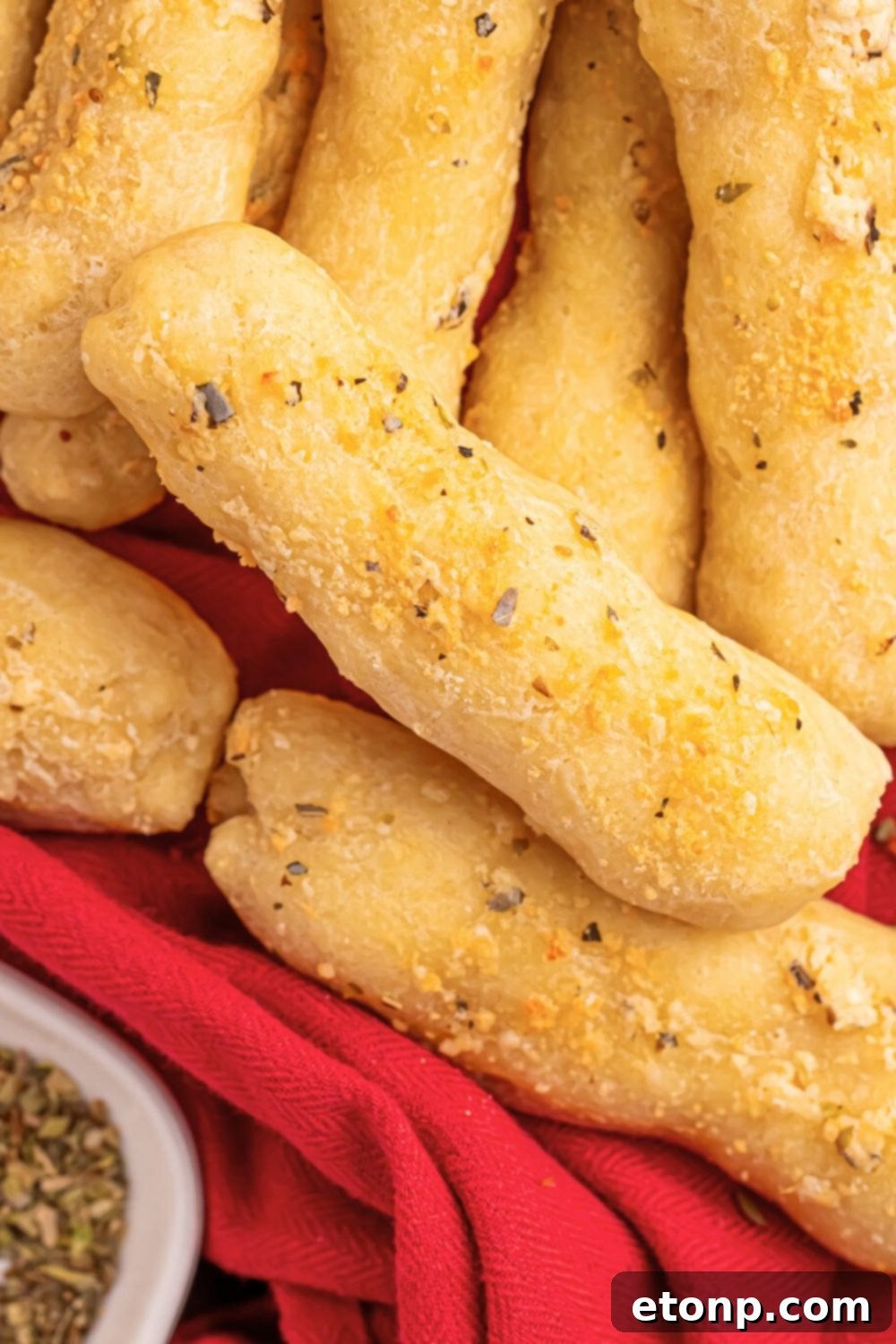 Easy Buttery Garlic Parmesan Breadsticks Two Ingredients 16 A stack of 2-Ingredient Garlic Parmesan Breadsticks resting on a red kitchen towel, with a small bowl of seasoning behind them on a wooden table.