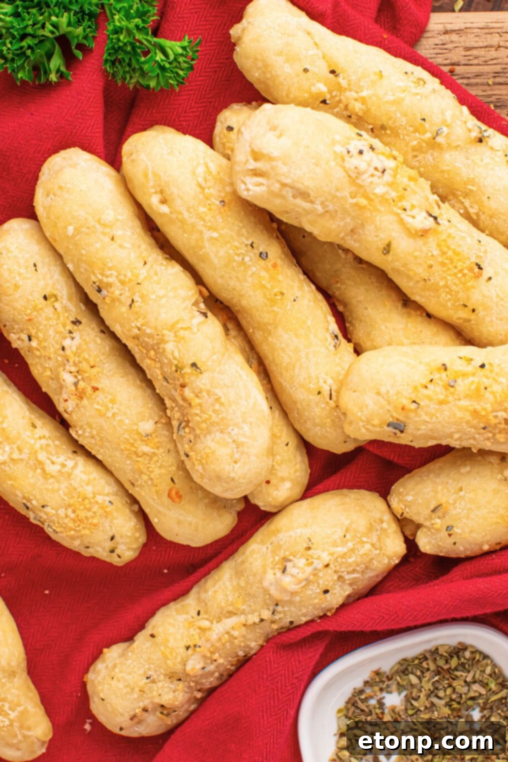 Easy Buttery Garlic Parmesan Breadsticks Two Ingredients 21 A red kitchen towel with a stack of 2-Ingredient Garlic Parmesan Breadsticks on top, and a sprig of parsley behind them on a wooden table.