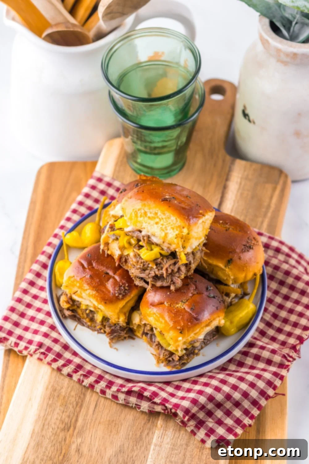 Slow Cooker Mississippi Pot Roast Sliders stacked on a plate with a towel under it with some glasses behind it on table 