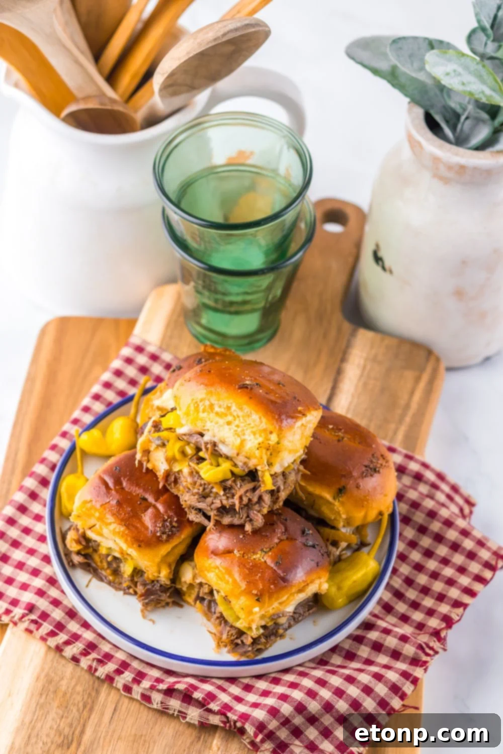Slow Cooker Mississippi Pot Roast Sliders on a table setting with glasses, cutting board and drink glasses behind it on table 
