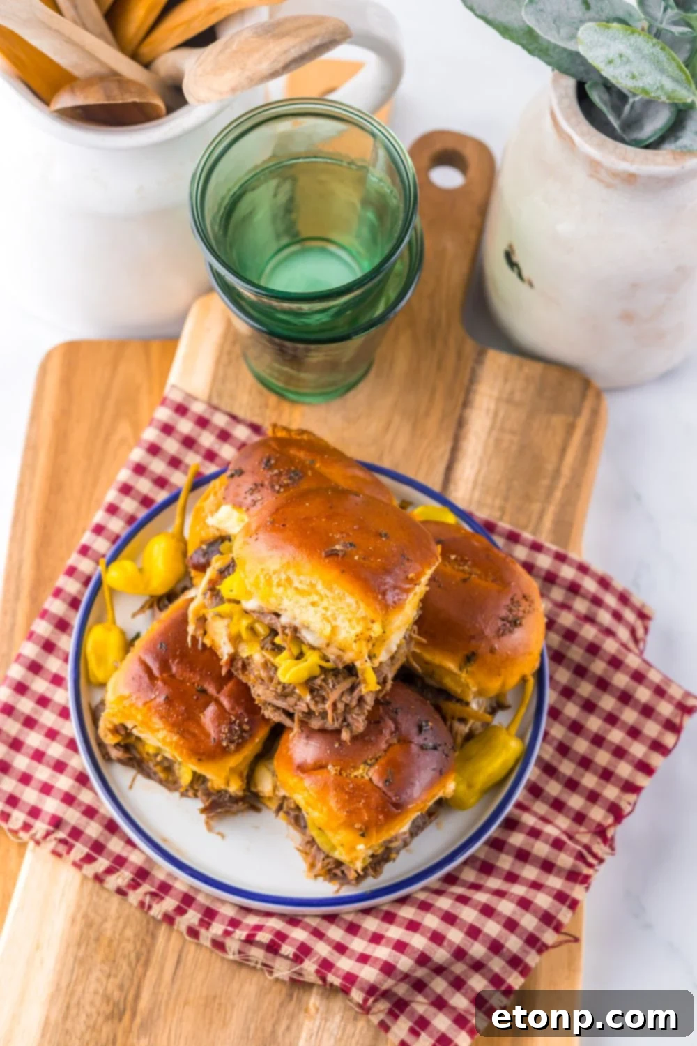 Slow Cooker Mississippi Pot Roast Sliders stacked on a plate with some glasses and ceramic containers behind it on table 