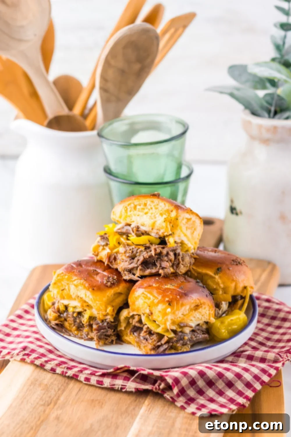 Slow Cooker Mississippi Pot Roast Sliders on a plate with drinks behind it and a jar full of wooden kitchen utensils 