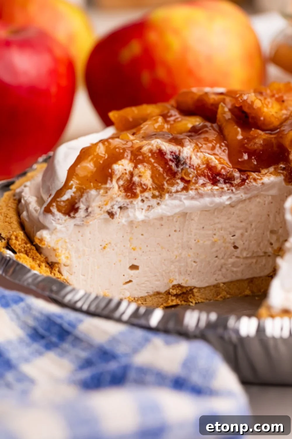 close up of No Bake Apple Cheesecake pie in pan with a slice taken out to see the layers of the cheesecake and apples in the background 