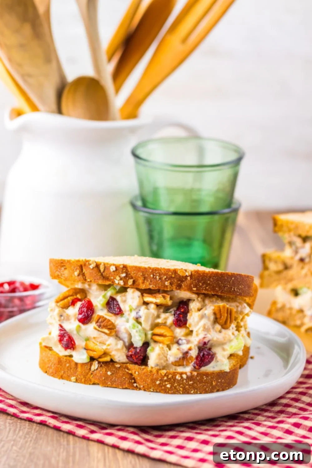 a thick turkey sandwich loaded with mayonnaise, celery, cranberries and more on a white plate on table with a checkered napkin spread out under the plate, with a rustic look to the table 