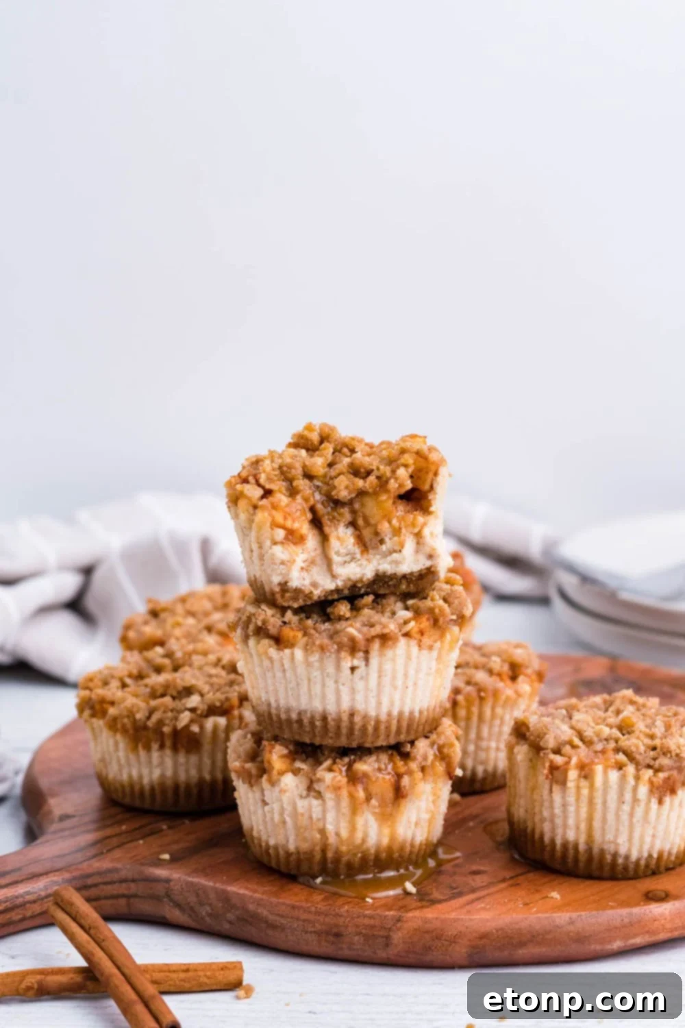 wooden cutting board sitting on marble counter with a towel behind it on table. On the tray is stacks of Apple Crisp Cheesecake Bites with one of the cheesecakes with some bites taken out of it 