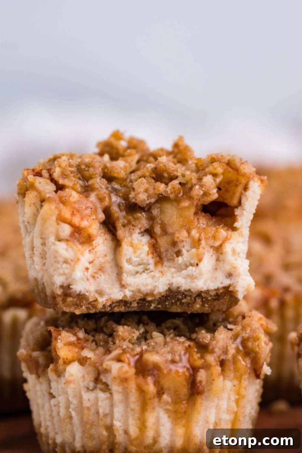 two Apple Crisp Cheesecake Bites stacked on top of each other on wooden cutting board. The top cheesecake has a few bites taken out of it 