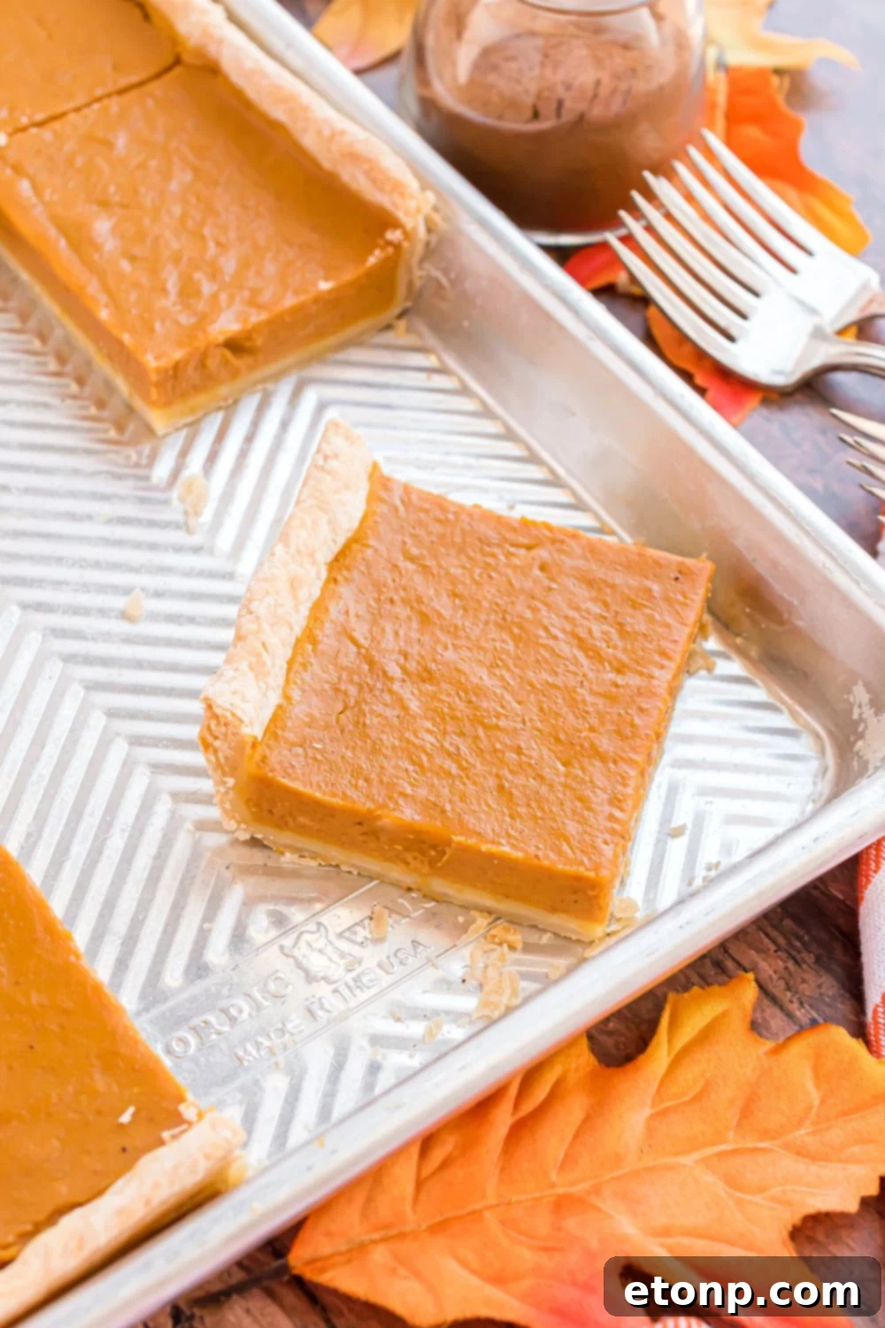 Effortless 4-Ingredient Sheet Pan Pumpkin Pie 18 metal pan on a wooden table decorated with rustic theme, with slices of Sheet Pan Pumpkin Pie inside the pan cut into squares