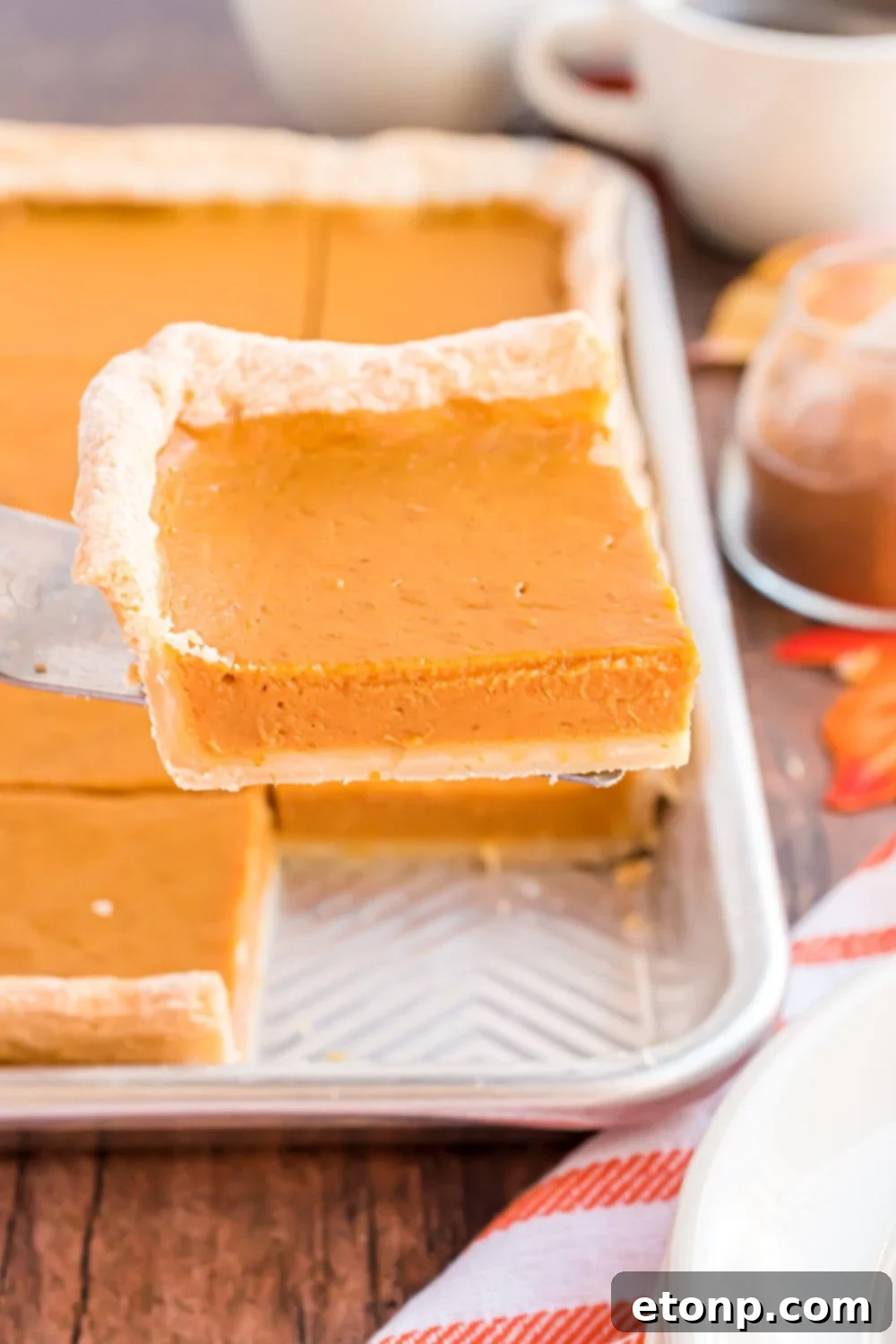 Effortless 4-Ingredient Sheet Pan Pumpkin Pie 19 Sheet Pan Pumpkin Pie in sheet pan on wooden table decorated for fall. Once slice of the pie beign lifted out of the tray to show the thickness