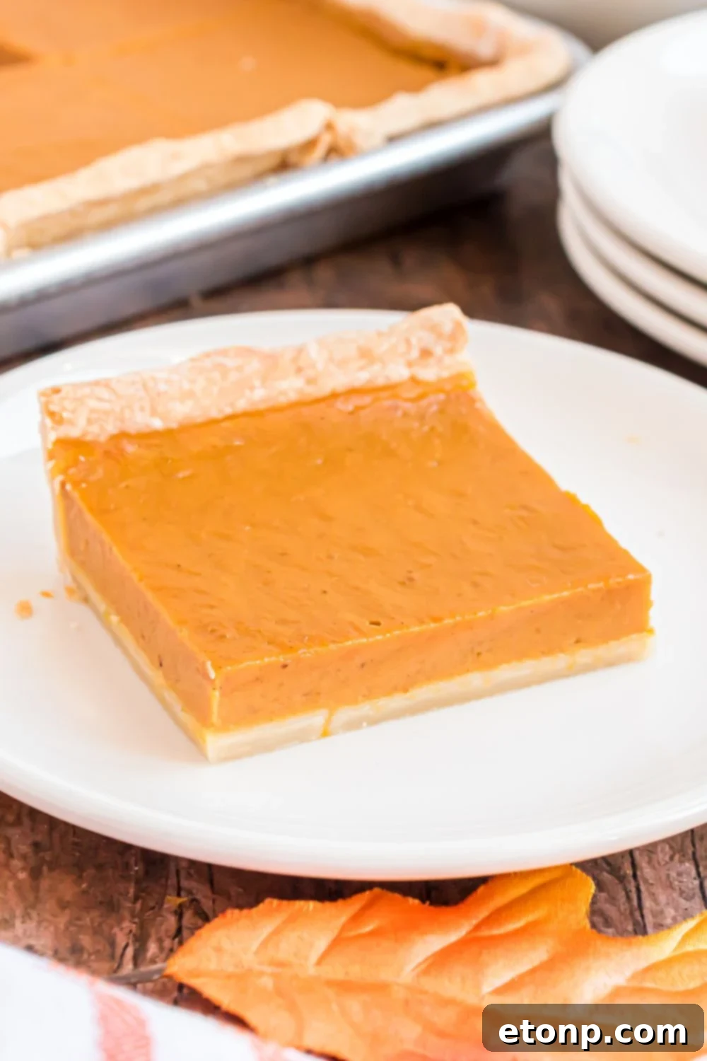 Effortless 4-Ingredient Sheet Pan Pumpkin Pie 23 slice of Sheet Pan Pumpkin Pie on a white plate with a stack of white plates to side on wooden table and sheet pan pie behind it on table
