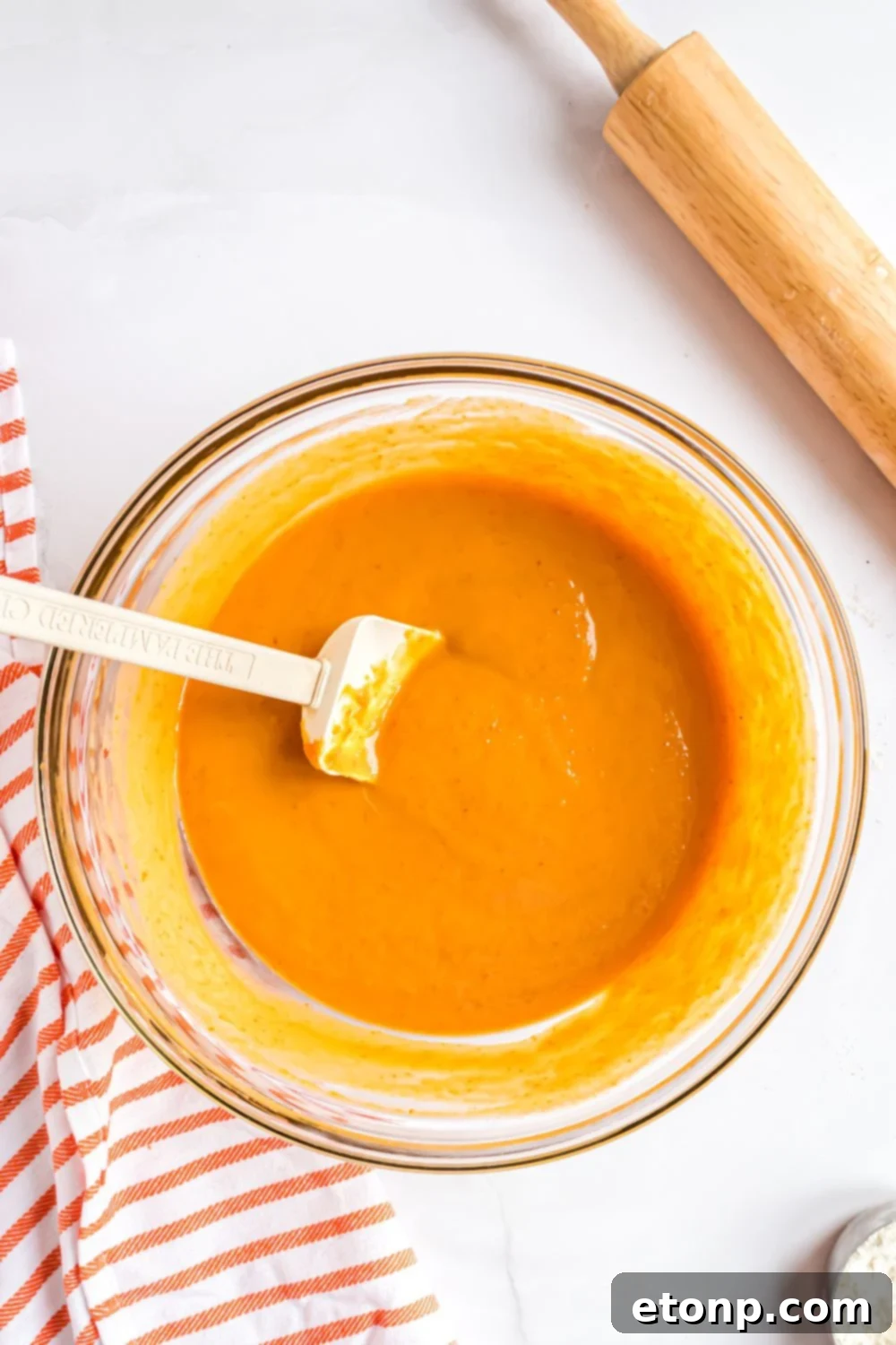 Effortless 4-Ingredient Sheet Pan Pumpkin Pie 9 glass bowl sitting on white table that has pumpkin pie batter fully mixed up with a red striped towel to side and rolling pin to the side
