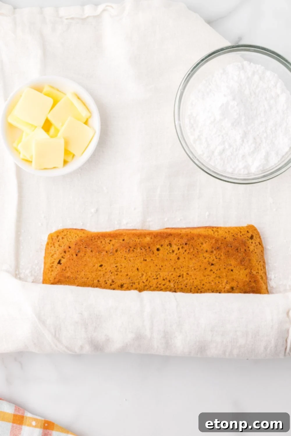 rolling up a Pumpkin Cake Roll with parchment paper. To the side are two small bowls one with sliced butter the other with powdered sugar