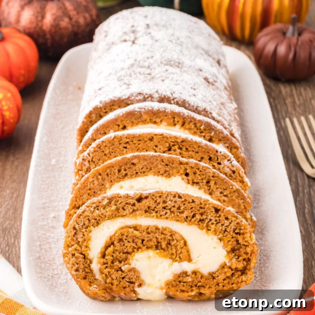 Pumpkin Roll on a long platter sitting on wooden table with pumpkins around to set a fall theme. Part of the pumpkin roll is sliced and the other part is not sliced and dusted with powdered sugar