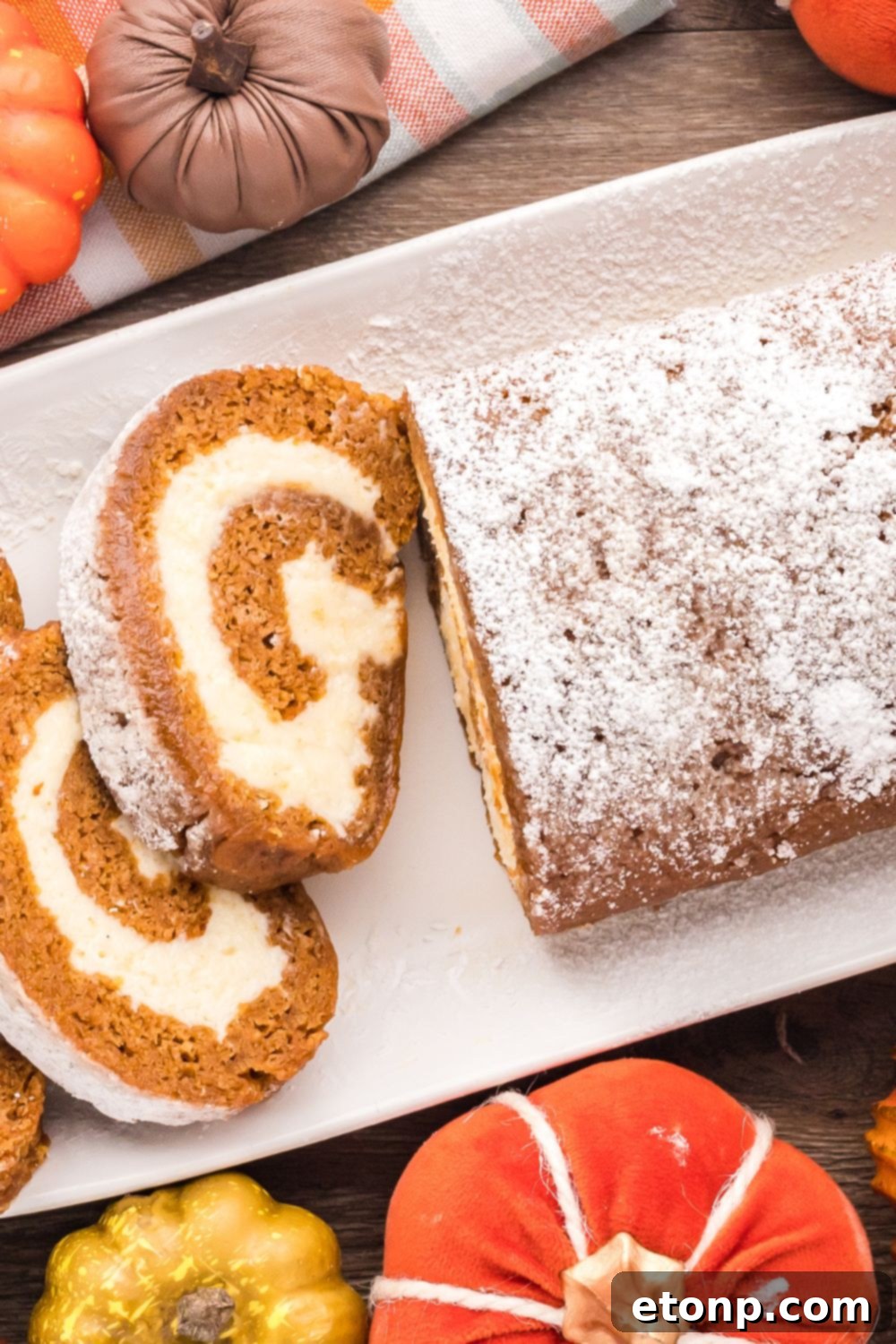 Pumpkin Cake Roll on a long white platter, with part of it whole and the other part sliced. On the wooden table around the platter are mini pumpkins for a rustic fall display