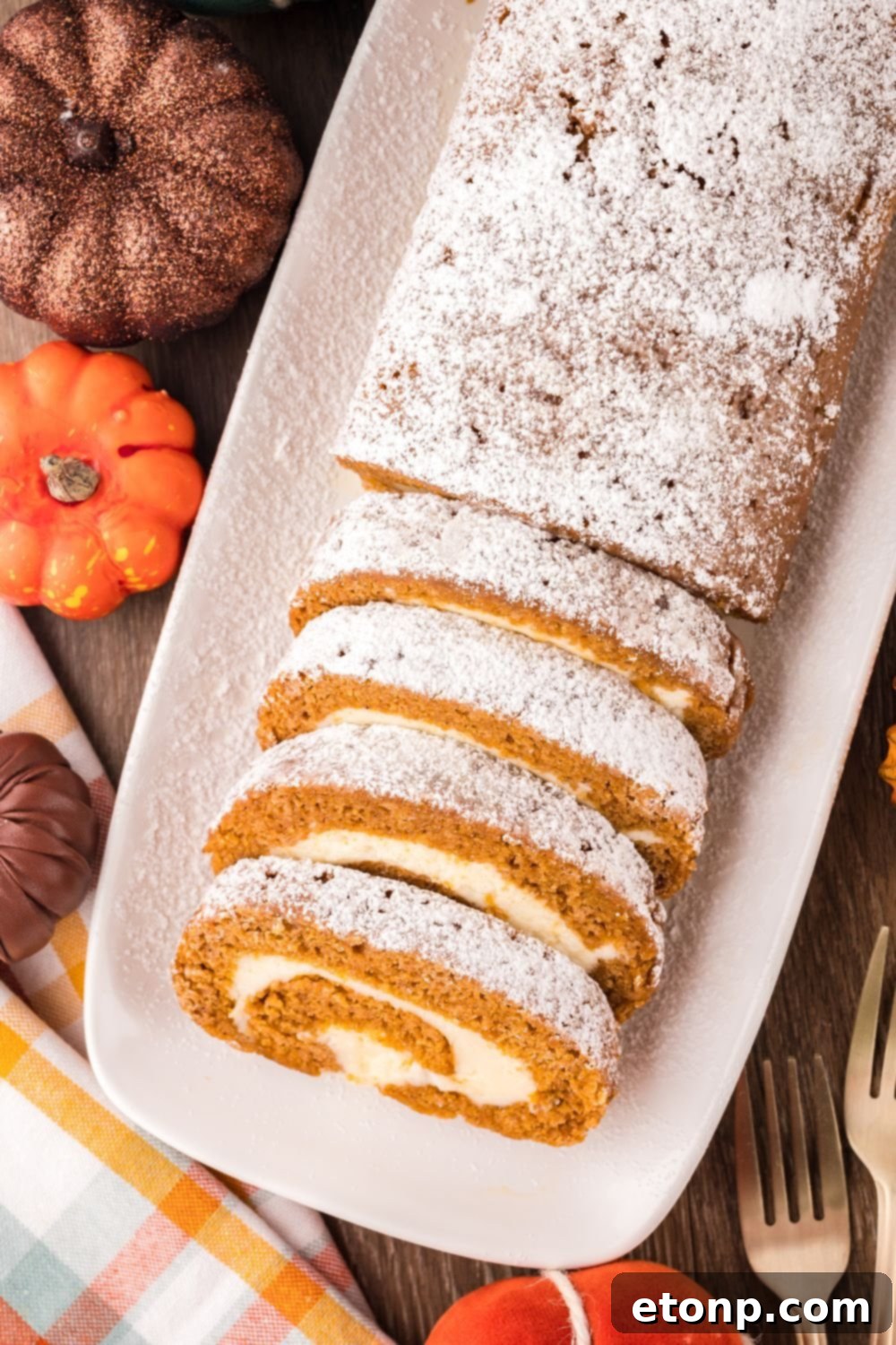 overhead look down on a wooden table with fake mini pumpkins and a long Pumpkin Cake Roll on a white platter. Part of the roll is sliced into wedges