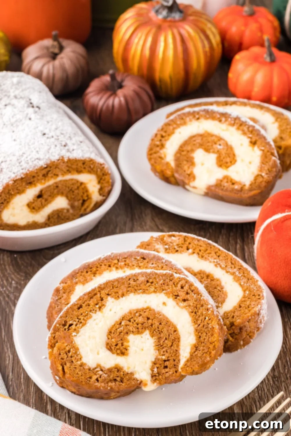 Pumpkin Cake Roll placed on plates, with a small platter to side with more of the pumpkin roll. Mini pumpkins scattered on table for a Thanksgiving fall look