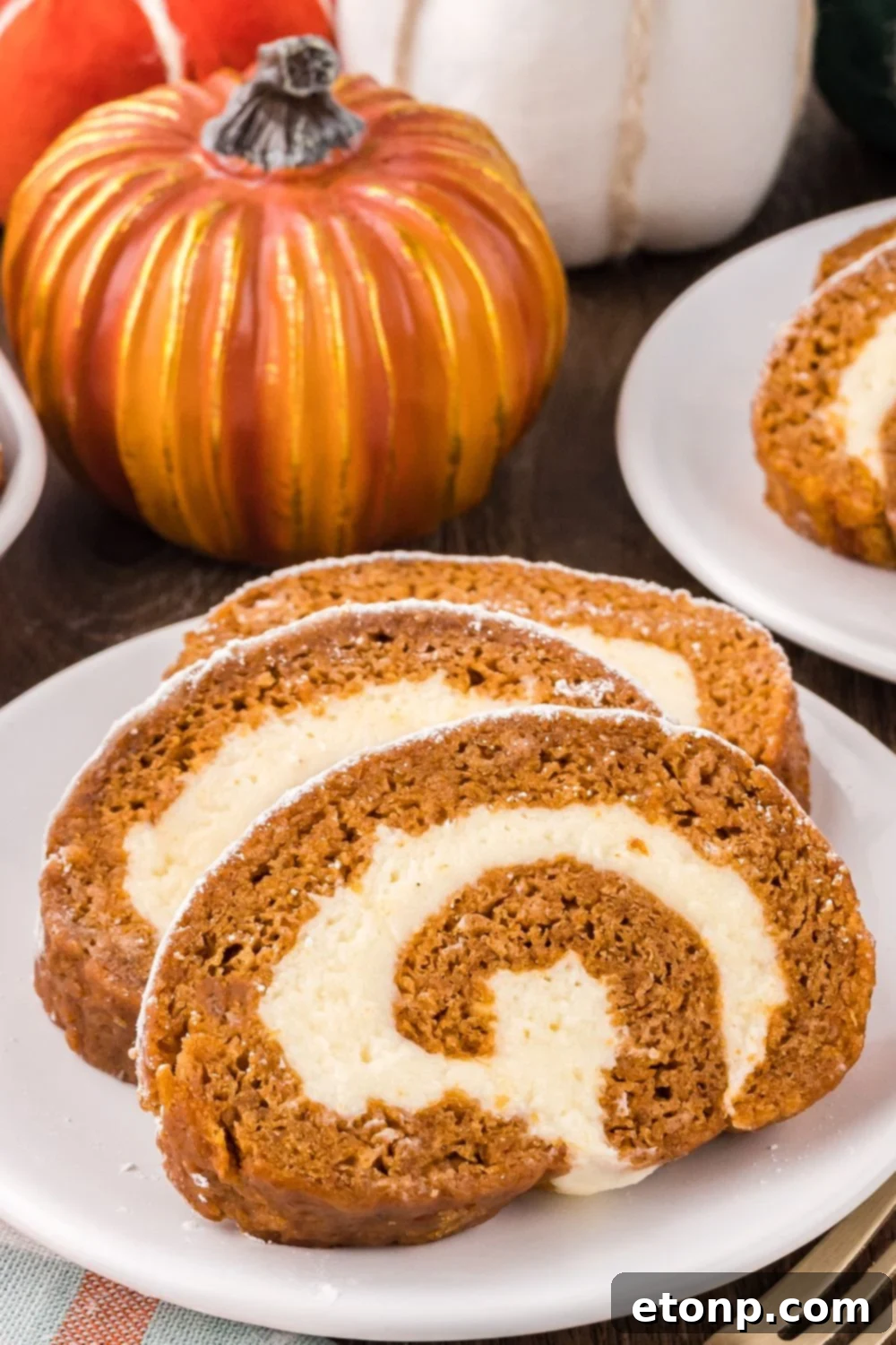 Pumpkin Cake Roll sliced on white plates placed on table with a few faux pumpkins around the plates to give a fall theme to table