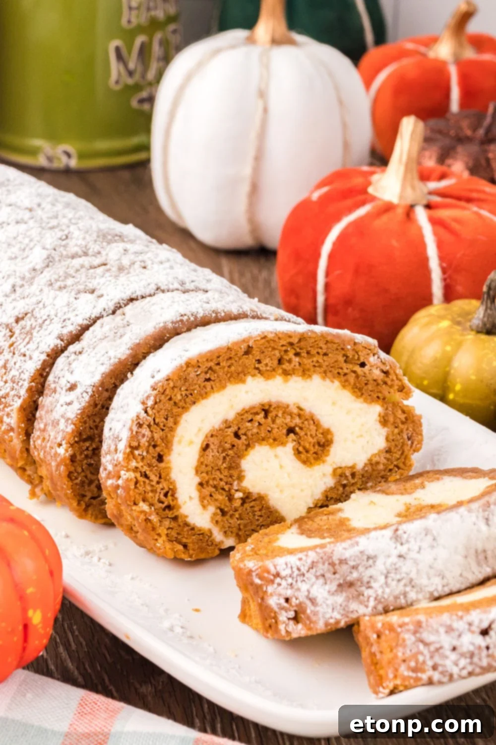 Pumpkin Cake Roll placed on table sitting on a white platter, with a portion of the pumpkin cake sliced. Then you have a mix of faux pumpkins scattered on table for a rustic fall look
