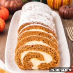 Pumpkin Roll on a long platter sitting on wooden table with pumpkins around to set a fall theme. Part of the pumpkin roll is sliced and the other part is not sliced and dusted with powdered sugar