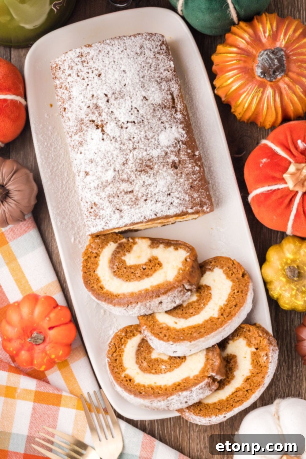 Pumpkin Cake Roll on a long white platter, with half of the pumpkin roll sliced. Faux pumpkins around the table with a white and brown striped towel