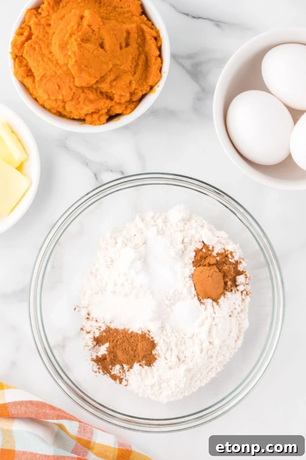 flour, spices and more in a glass bowl with small bowls around it with eggs, pumpkin and sliced butter for making Pumpkin Cake Roll