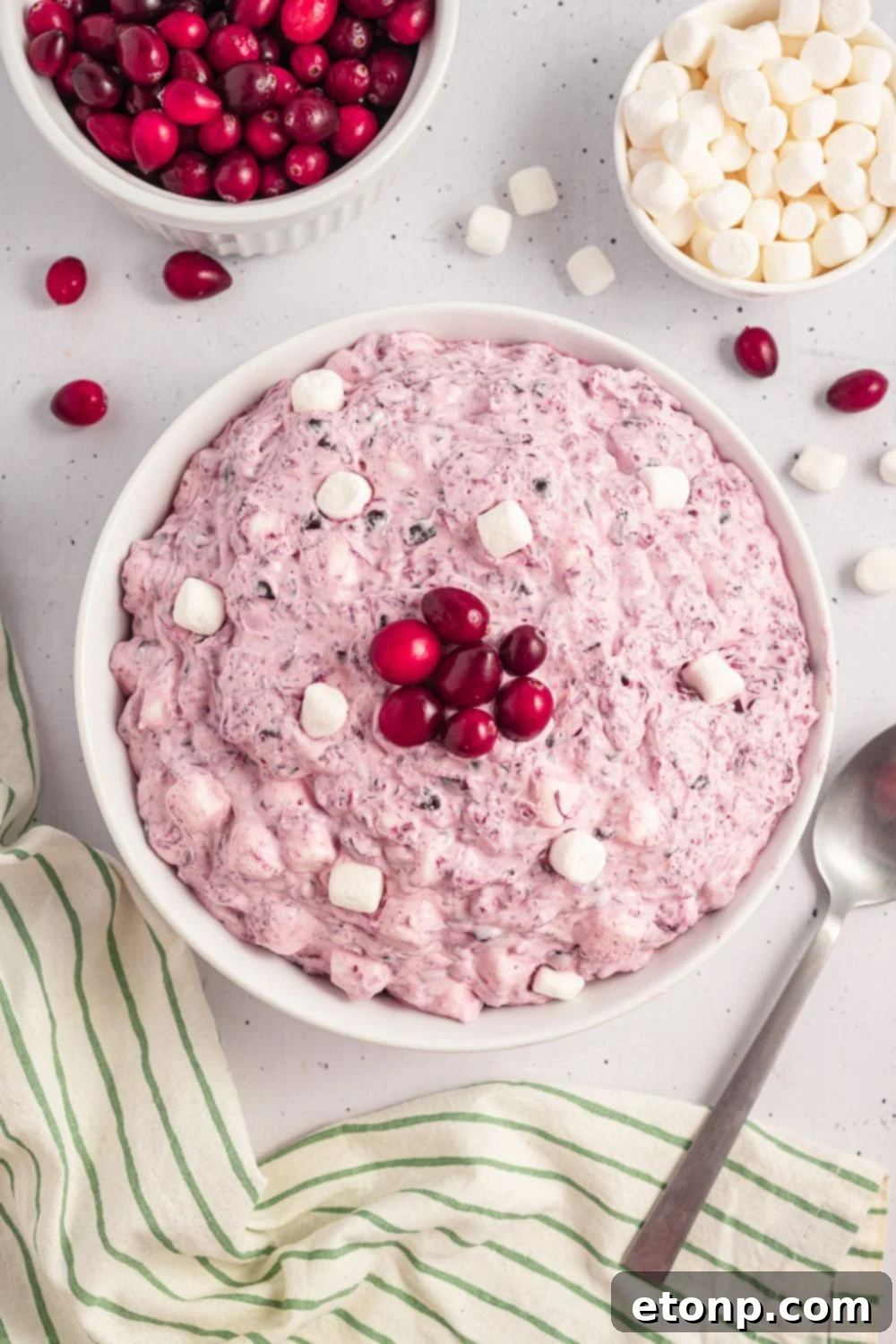 white table with a large bowl of Cranberry Fluff Salad, a spoon to side of bowl. Then you have mini marshmallows in a bowl, cranberries, and a few scattered around table 