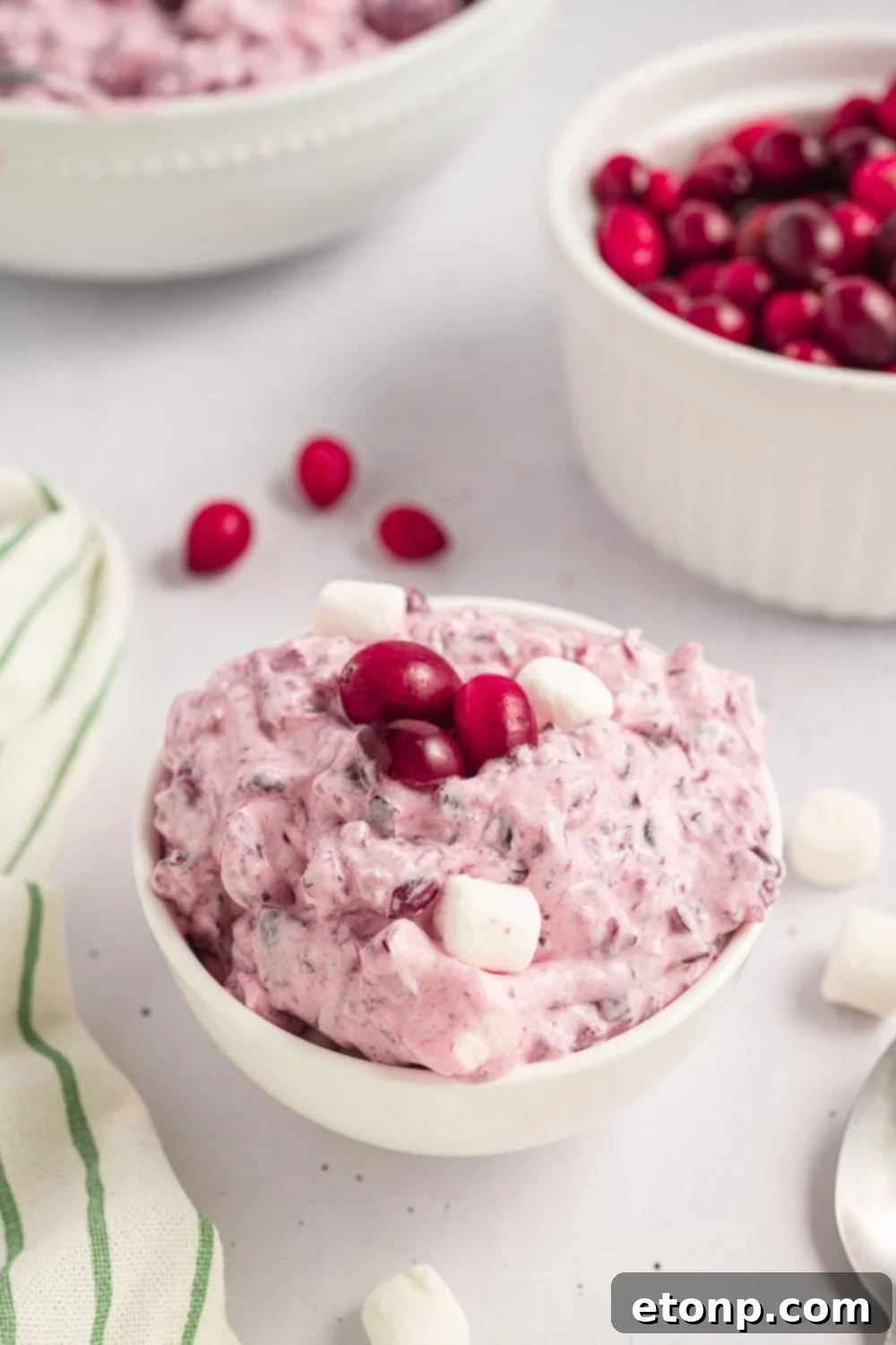 Cranberry Fluff Salad in a white bowls on table with a bowl of cranberries in another bowl, and a striped towel to side of table 