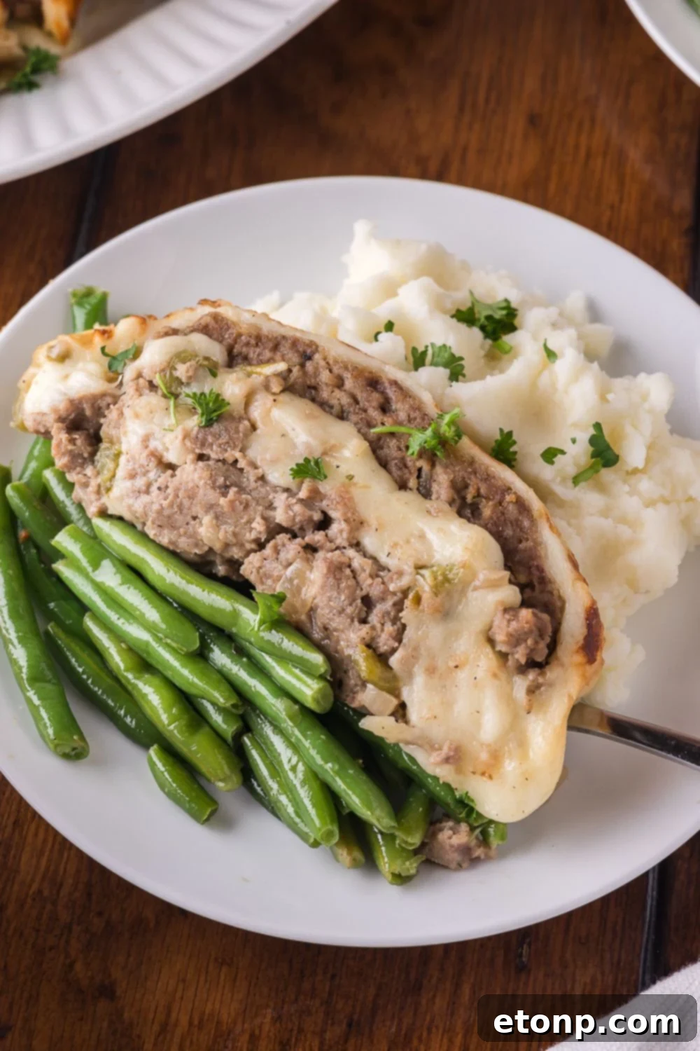 Cheesy Philly Stuffed Meatloaf with Peppers and Onions 14 wooden table with a white plate sitting on it, that has green beans, philly cheesesteak meatloaf, and a spoonful of mashed potatoes