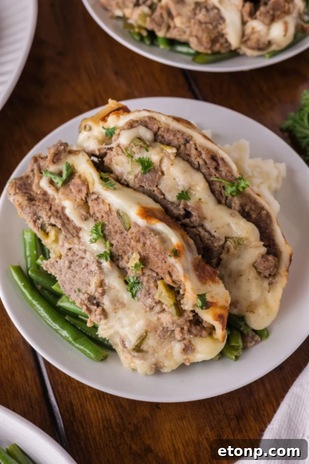 Cheesy Philly Stuffed Meatloaf with Peppers and Onions 15 two plates on table with some green beans and large slices of Stuffed Philly Cheesesteak Meatloaf on top with mashed potatoes as well.