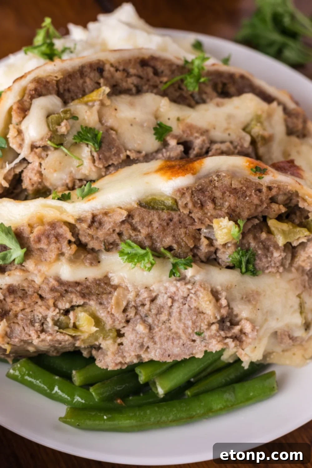 Cheesy Philly Stuffed Meatloaf with Peppers and Onions 16 white plate sitting on table with a bed of green beans, and on top thick slices of Stuffed Philly Cheesesteak Meatloaf topped with fresh parsley