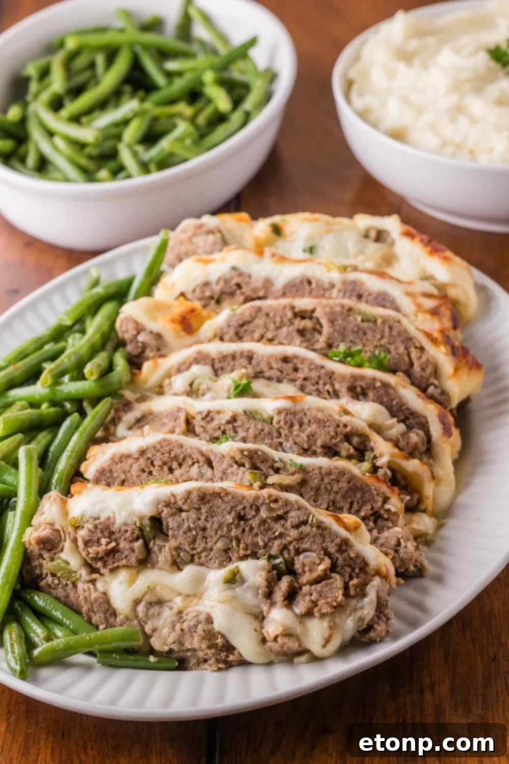 Cheesy Philly Stuffed Meatloaf with Peppers and Onions 19 wooden table with a bowl of green beans, bowl of mashed potatoes, and a platter with sliced Stuffed Philly Cheesesteak Meatloaf on platter with green beans on side