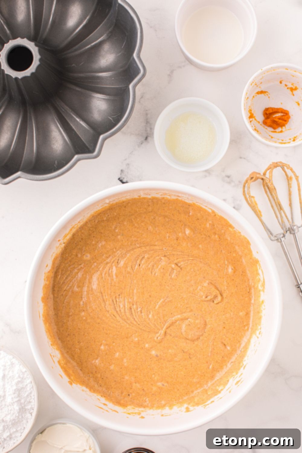 white bowl on table full of cake batter and a whisk to side and bundt pan