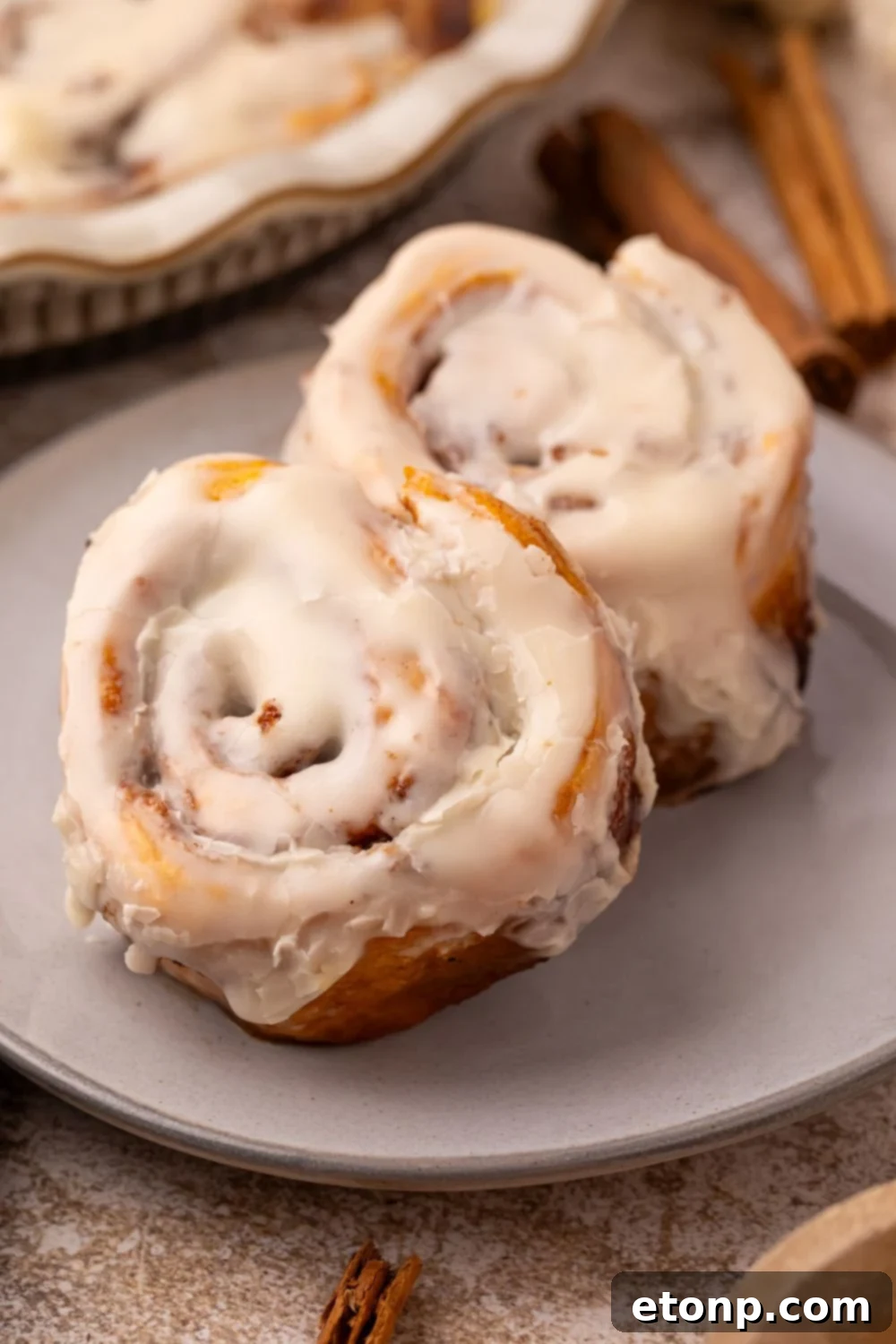 Effortless 2-Ingredient Pumpkin Cinnamon Rolls No Yeast 17 2 Pumpkin Cinnamon Rolls on a grey plate on table with a pan with more rolls behind it and a couple of cinnamon sticks on table