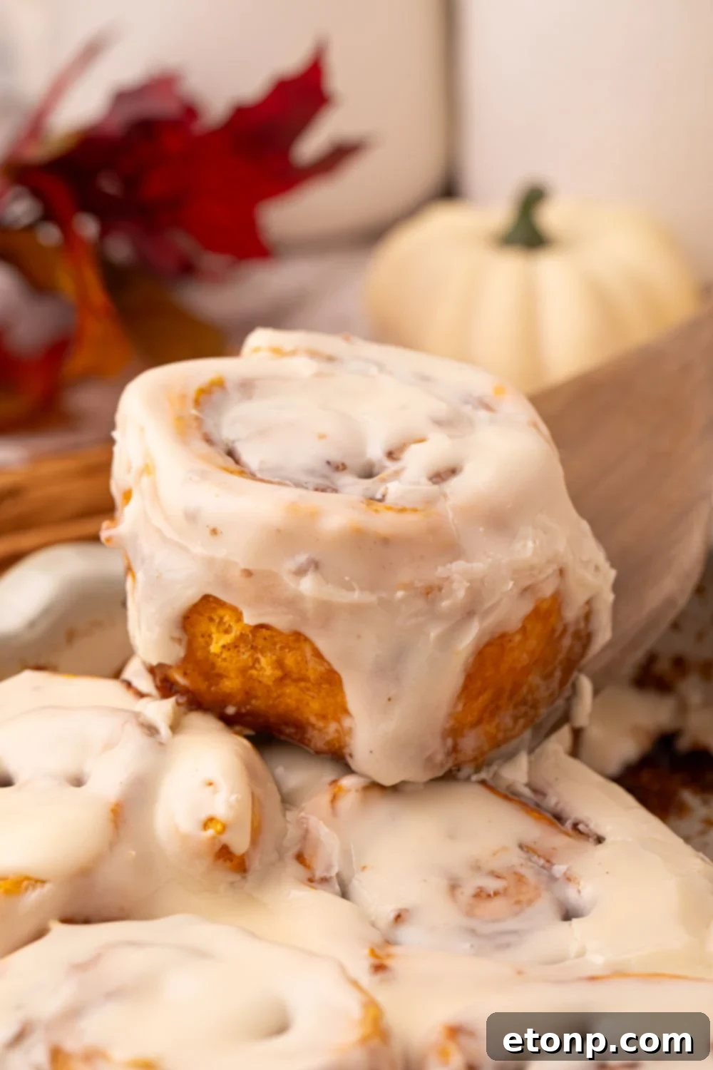 Effortless 2-Ingredient Pumpkin Cinnamon Rolls No Yeast 21 Pumpkin Cinnamon Roll being lifted out of container with a wooden spoon. The back of the image is decorated with fall items like leaves, pumpkins, etc