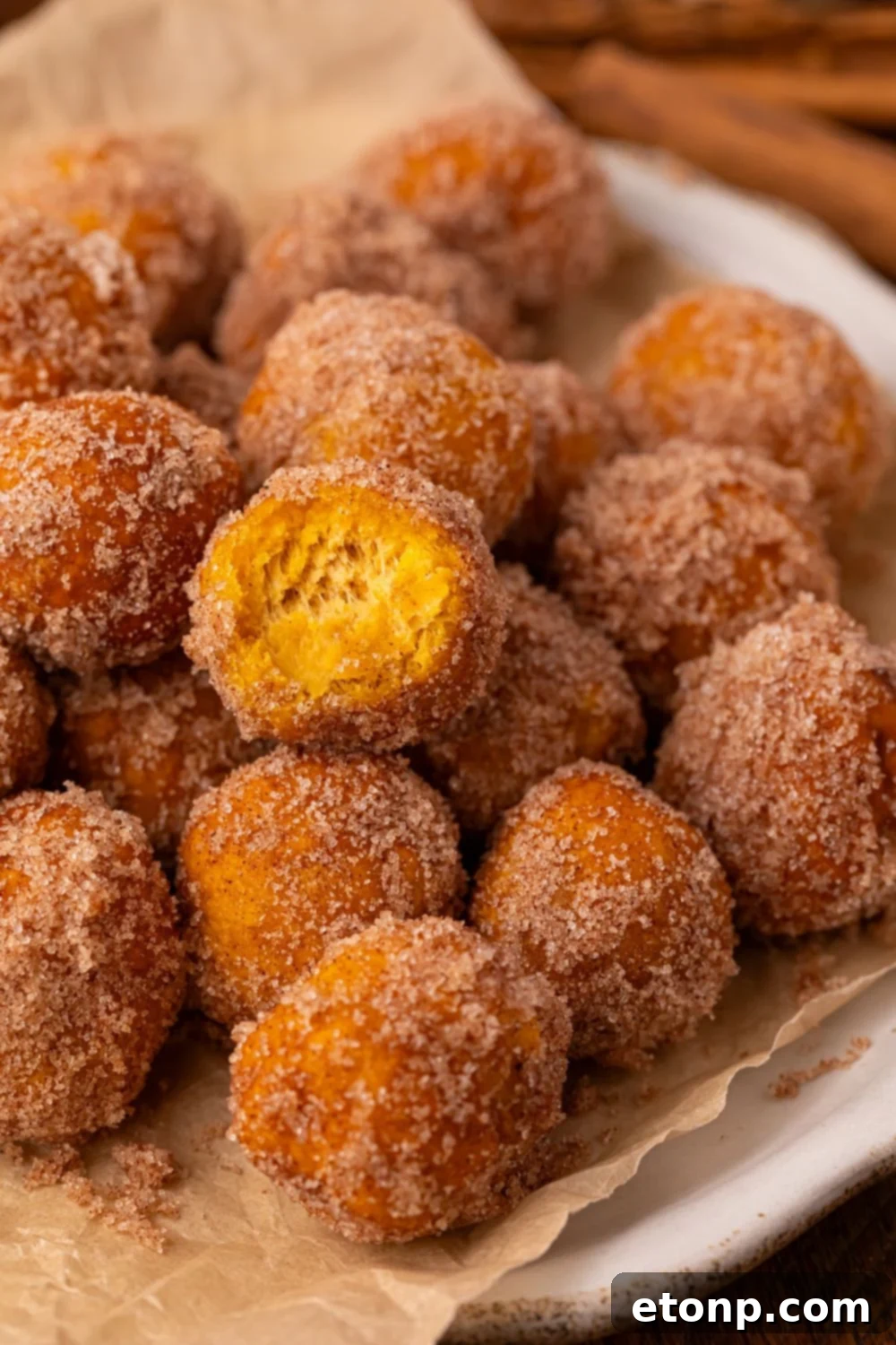 Effortless Two-Ingredient Pumpkin Pie Bites 12 Pumpkin Pie Donut Holes scattered on a parchment lined white platter on a wooden table. The top donut hole has a bite taken out of it.