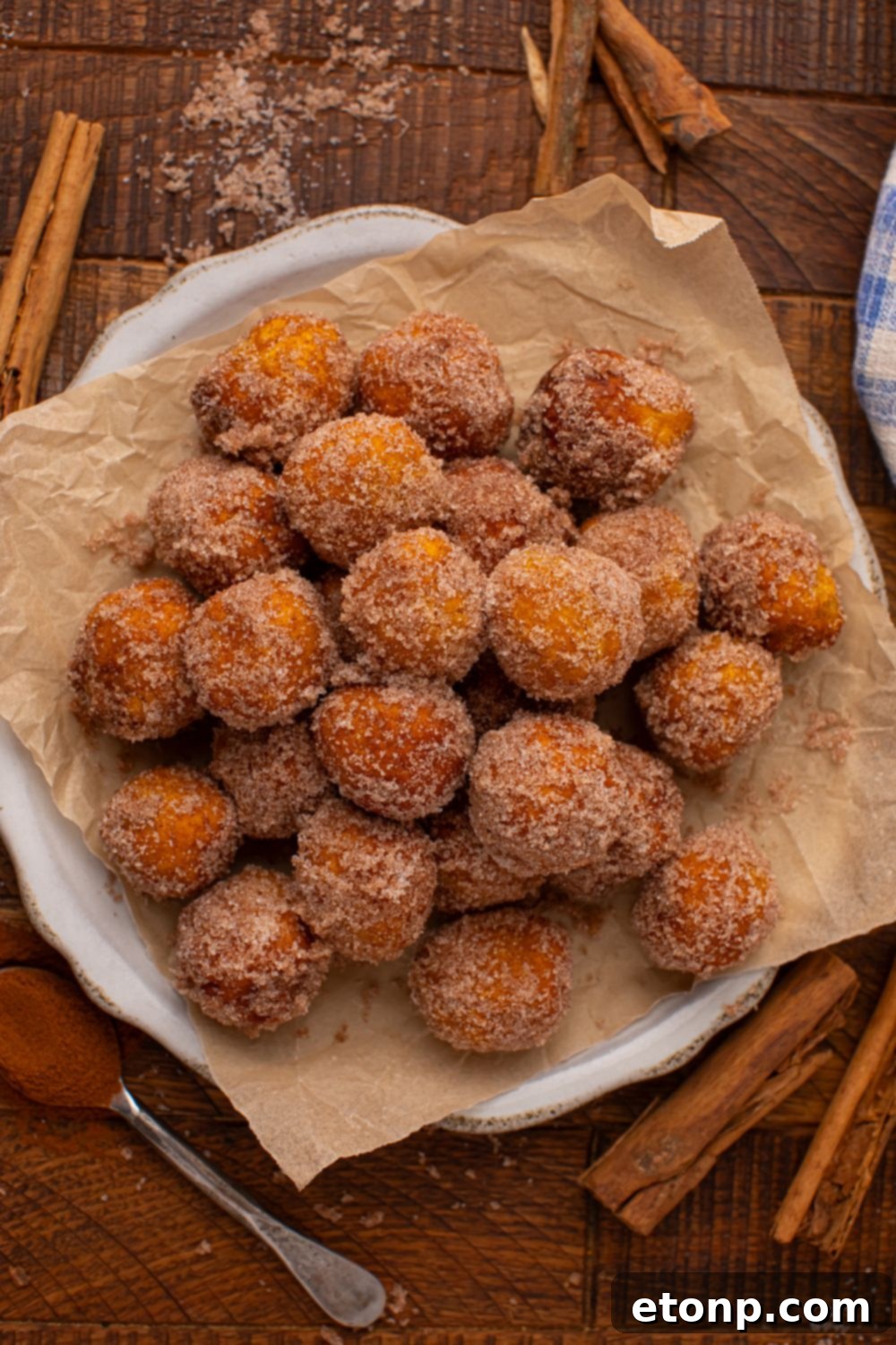 Effortless Two-Ingredient Pumpkin Pie Bites 21 Pumpkin Pie Donut Holes on a parchment lined white platter on wooden table. There are cinnamon sticks scattered around the table