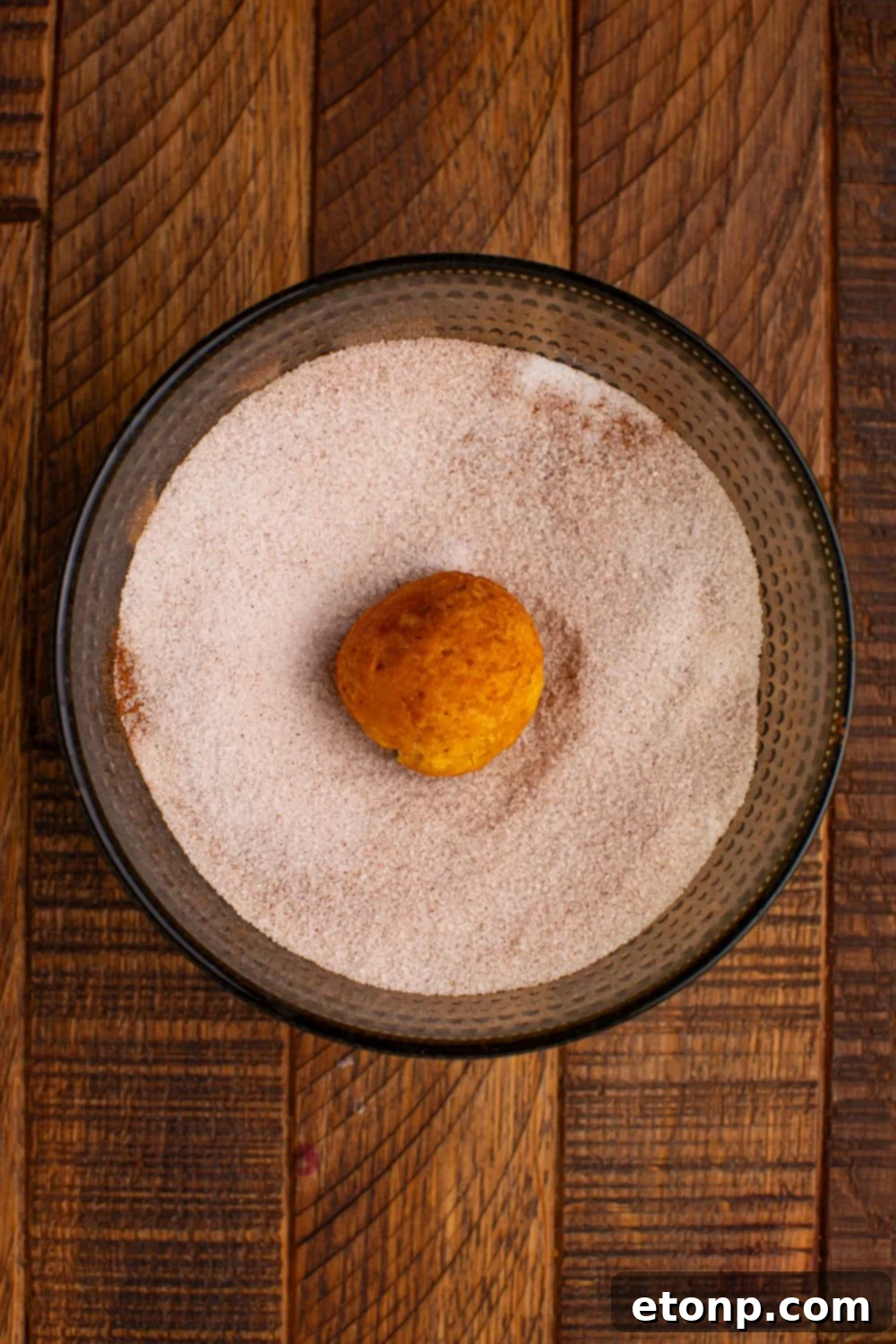 Effortless Two-Ingredient Pumpkin Pie Bites 6 cinnamon sugar mixture in a glass bowl with a donut hole in center of the bowl on a wooden table