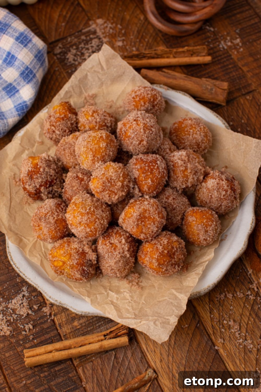 Effortless Two-Ingredient Pumpkin Pie Bites 9 wooden table with a white platter full of Pumpkin Pie Donut Holes. Cinnamon sticks and a blue checkered towel to side of bowl