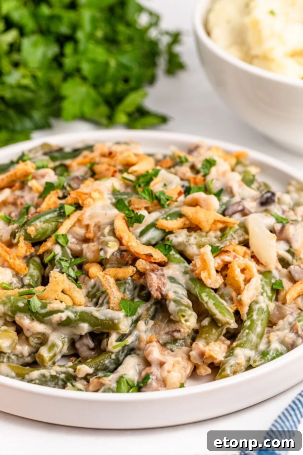 side view of a white plate with an edge sitting on table full of Green Bean Casserole and behind it on table fresh parsley bundle 