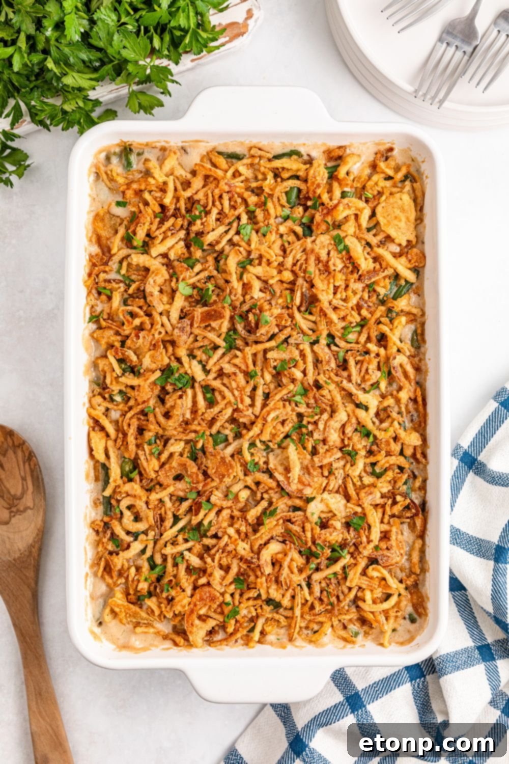 looking down on table to see a casserole dish with golden brown Green Bean Casserole and a wooden spoon to side, table napkin and fresh parsley 