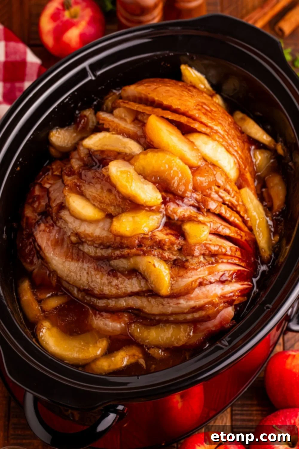 looking down at wooden table with a black slow cooker with apple glazed spiral ham. Fresh apples around table 