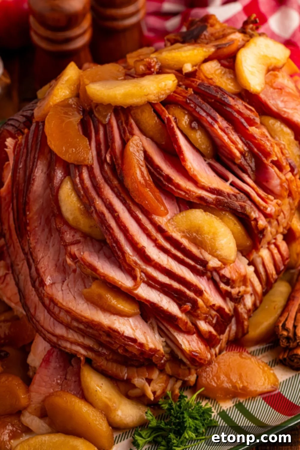 view of a large sliced spiral ham on platter with some apples on top and around with a red checkered towel behind it 