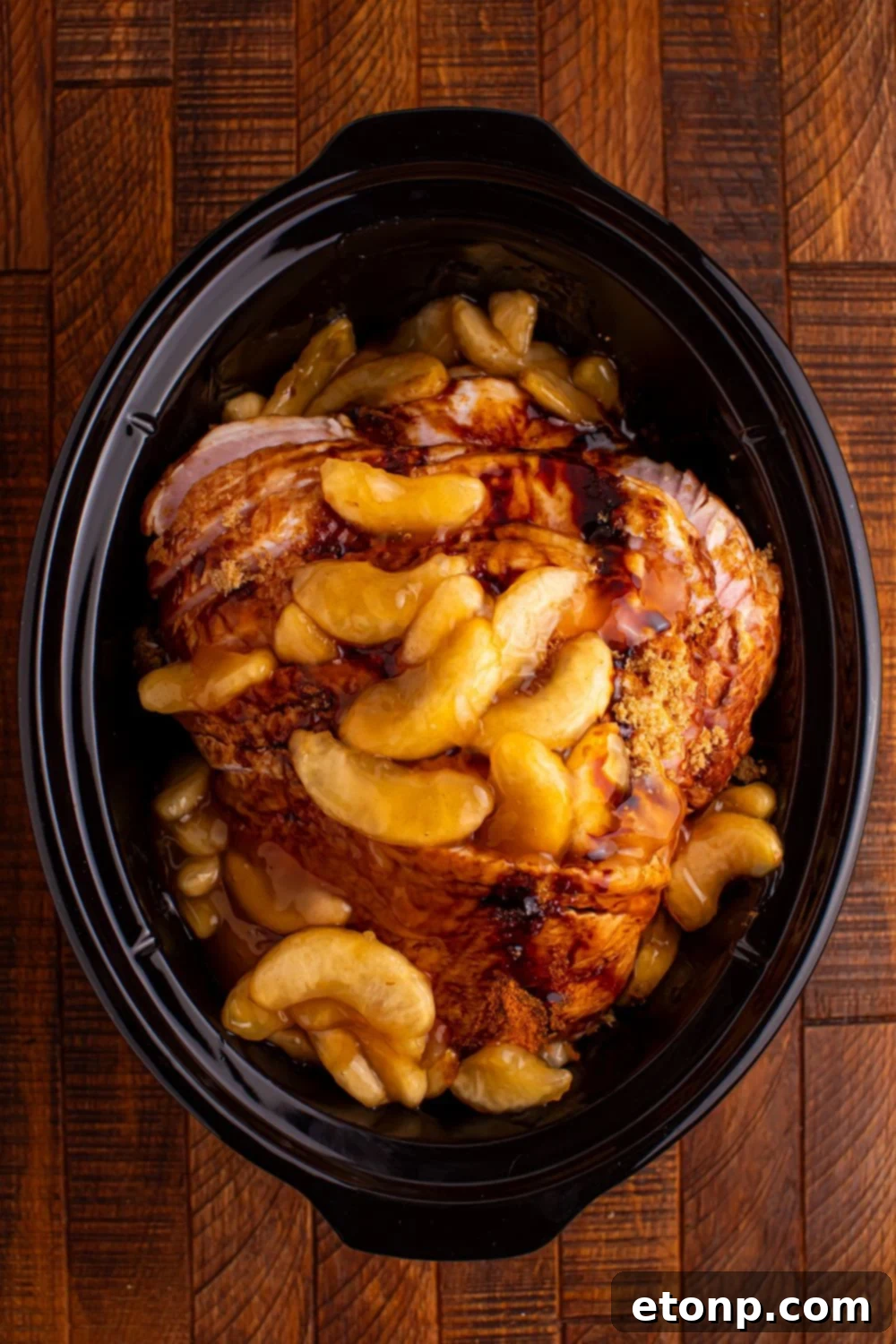 wooden table with a black crockpot in center with a sliced ham topped with spiced apples 