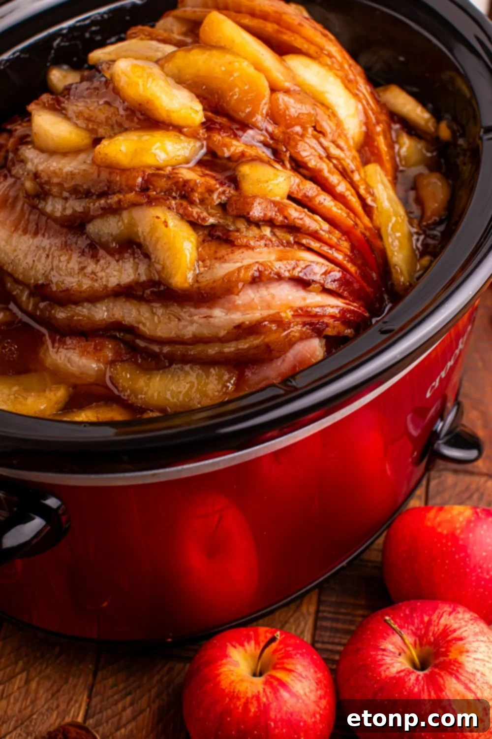 red slow cooker on a wooden table with a spiral ham with apples on top, and whole apples in front of table