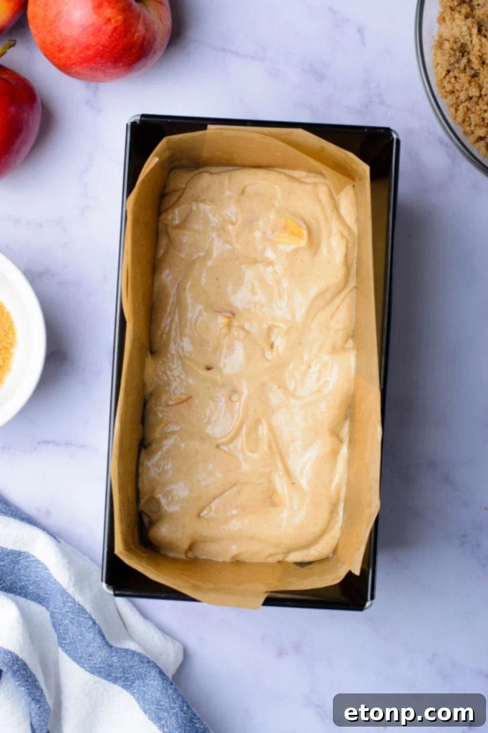 looking down at marble counter with a loaf pan with half full of a cinnamon loaf cake batter and apples and a brown sugar mixture to the side