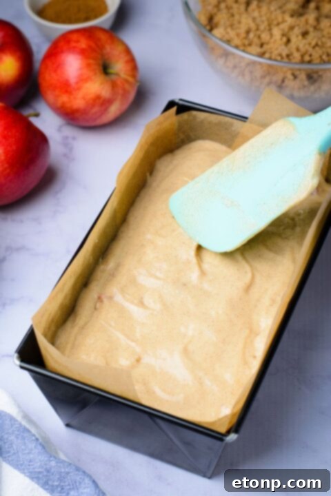 raw batter in a loaf pan being spread around with a spatula, and two apples to side of pan