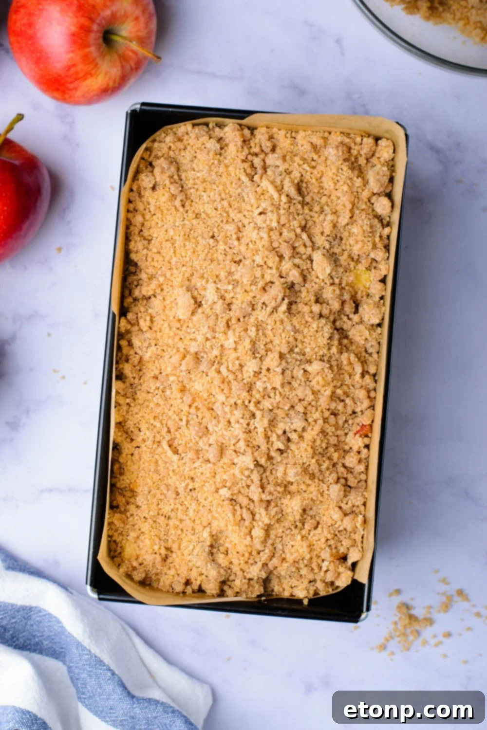 cinnamon crumble on top of a loaf cake batter in pan with a couple apples around the table
