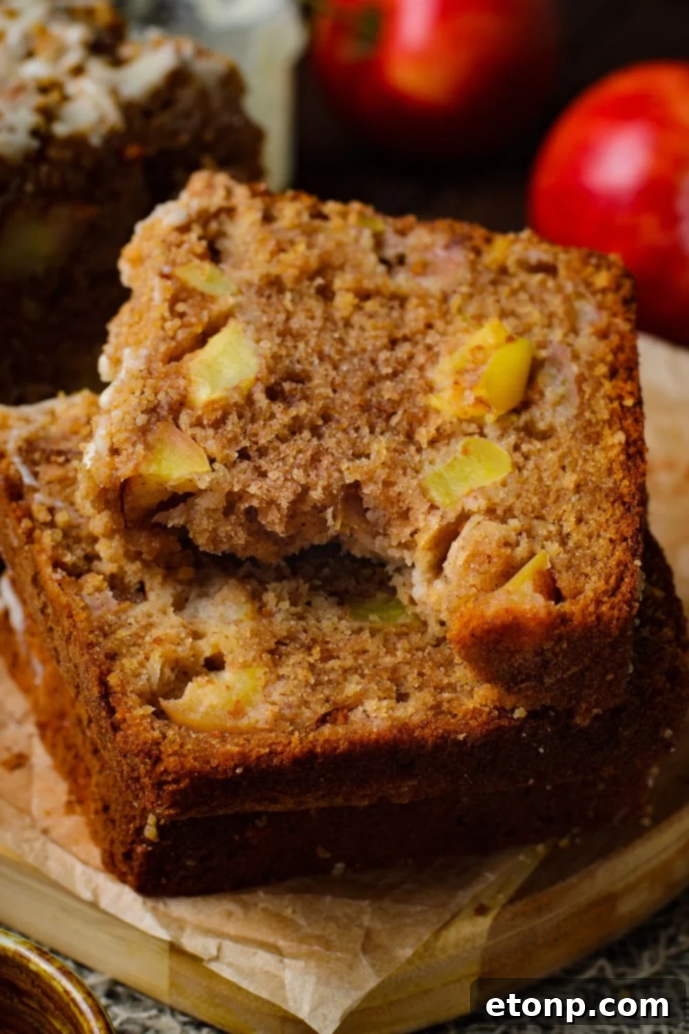 Apple Cinnamon Loaf Cake sliced into thick pieces stacked on top of one another on a tray. The top slice has a bite taken out, and red apples are behind it on table for a nice red color to image